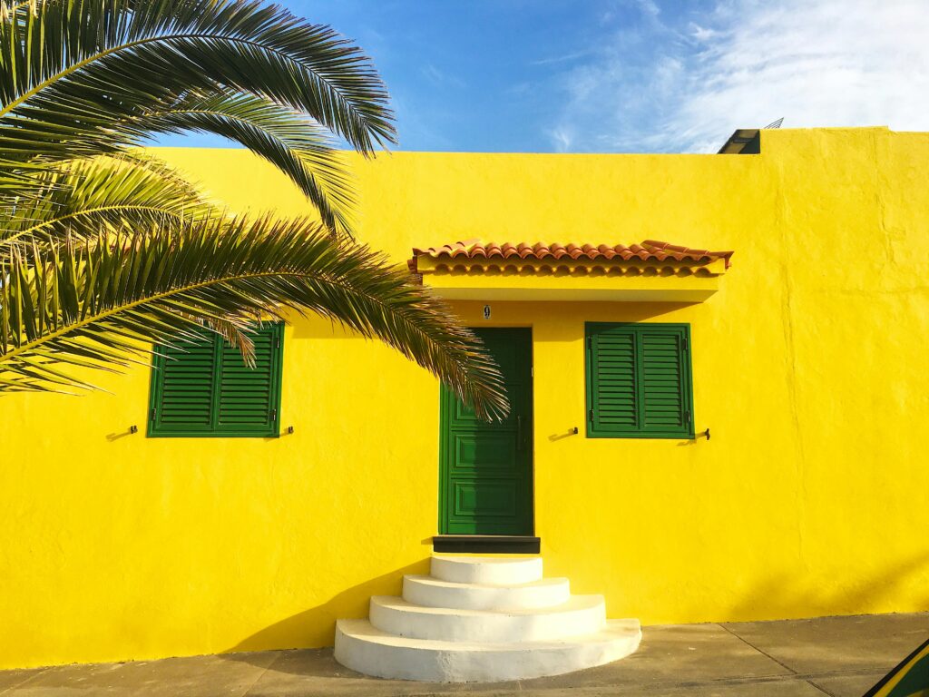 pexels-photo-12657392-12657392 Vibrant yellow house facade in Spain with green shutters and a palm tree.